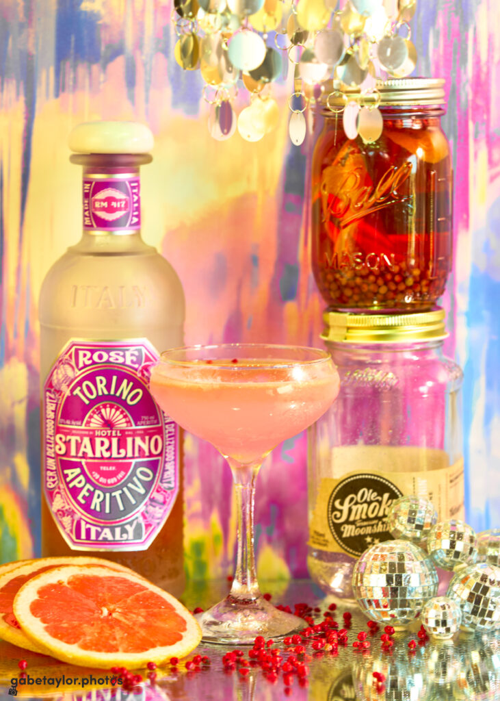 a pink cocktail in a coupe glass is surrounded by grapefruit slices, a bottle of Hotel Starlino Rosé Aperitivo, a mason jar filled with deep pink liquid, peppercorns, and grapefruit, a jar of Ole Smoky Moonshine, and miniature disco balls and scattered pink peppercorns